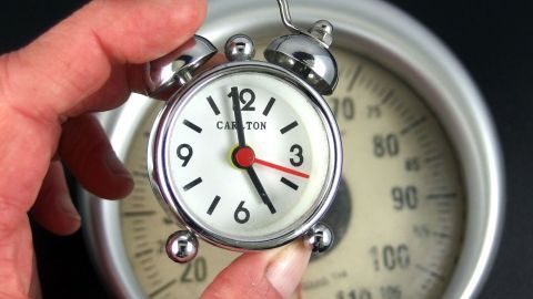 Eine Hand hält einen Wecker, während er auf eine Waage im Hintergrund zeigt, was die 17 Uhr Diät symbolisiert.