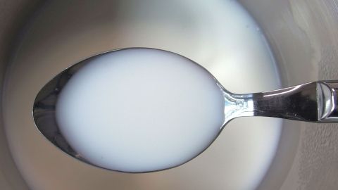 Eine silberne Löffelhalb mit lauwarmer Milch befindet sich in einer Schüssel, umgeben von leuchtend hellem Flüssigkeitsreflex.