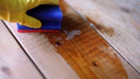 Eine Hand mit einem gelben Handschuh reinigt einen Holzterrassendielen mit einem Schwamm, um den Grünbelag zu entfernen.