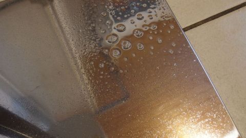 Ein glänzendes Edelstahlstück ist mit Fensterreiniger besprüht, während Wassertropfen darauf sichtbar sind.