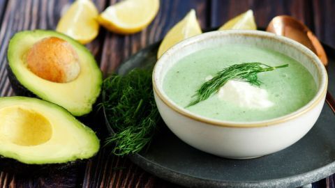 In einer hellen Schüssel wird eine cremige Avocadosuppe mit frischem Dill serviert, umgeben von geschnittenen Avocados und Zitronen.