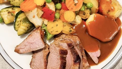 Ein Teller mit zartem Fleisch, umgeben von buntem Gemüse, Kartoffeln und einer dunklen Soße zum Fleisch.