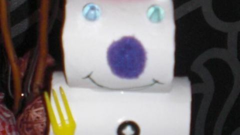 Ein Schneemann aus Klopapierrollen lächelt mit buntem Hut und gelbem Besteck in einem festlich dekorierten Raum.