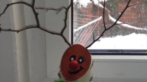 Ein Rentier aus einem Baumstamm steht mit Astgeweihen und einem lächelnden Gesicht auf einem Fensterbrett in einer verschneiten Umgebung.