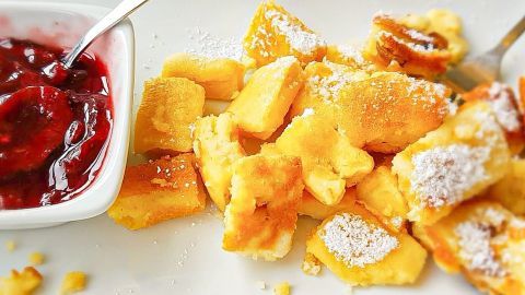 Kaiserschmarrn-Stücke liegen auf einem Teller, bestäubt mit Puderzucker, neben einem Becher mit fruchtiger Sauce.