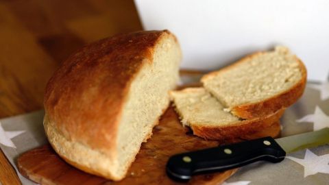 Ein frisch gebackenes Brot liegt auf einem Holzbrett, während ein Stück davon mit einem schwarzen Messer geschnitten wurde.
