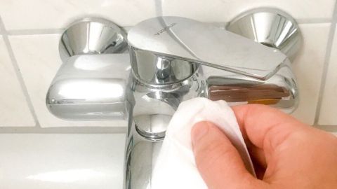 Eine Hand hält ein Reinigungstuch und wischt über einen glänzenden Wasserhahn in einem Badezimmer.