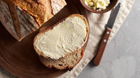 Eine Scheibe Brot ist mit Rapsbutter bestrichen und liegt auf einem Holzbrett, umgeben von einer Schüssel mit Rapsbutter.