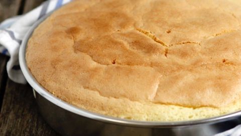 Ein runder Biskuit liegt frisch gebacken in einer silbernen Backform, umgeben von einem blauen Küchentuch auf einem Holztisch.