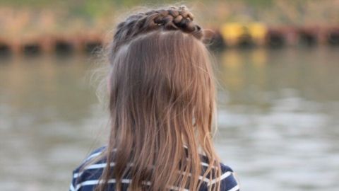 Ein kleines Mädchen mit langen, geflochtenen Haaren steht am Ufer eines Gewässers und betrachtet das Wasser.