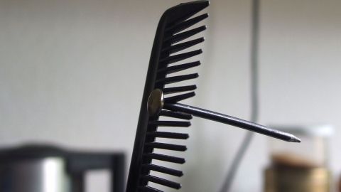 Nagel in einen Kamm "einspannen" und damit festhalten - so kann man sich nicht auf die Finger hämmern. Ein Kamm steht senkrecht, während ein Nagel darin steckt, auf einem Tisch in einem gemütlichen Raum.