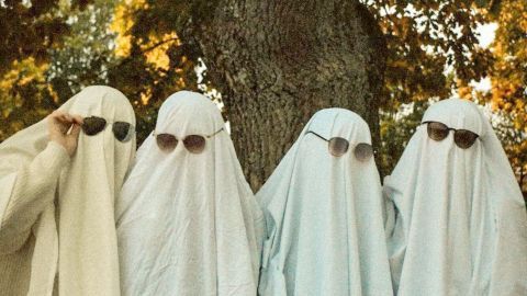 Vier Geister in simplen Karnevalskostümen tragen Sonnenbrillen, posierend vor einem Baum im Freien bei Sonnenuntergang.