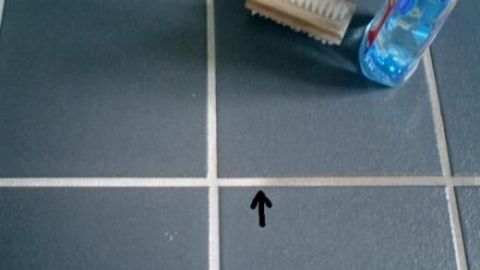 Eine Fliesenfuge zeigt Verschmutzung, während eine Bürste und eine Reinigungsflasche daneben auf dem Boden liegen, bereit zur Reinigung.