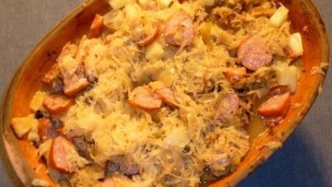 Ein Römertopf enthält frisch zubereitetes Sauerkraut mit Würstchen und Kartoffeln, serviert auf einem blauen Tisch.