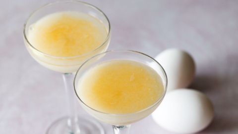 Zwei elegante Cocktails mit Eierlikör stehen auf einem hellen Tisch, neben frischen Eiern und einem grünen Blatt.
