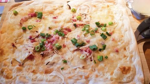 Flammkuchen auf Holzbrett, belegt mit Zwiebeln, Speck und Frühlingszwiebeln, in einer warmen Küchenszene angerichtet.
