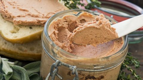 Eine cremige Leberwurst wird mit einem Spatel aus einem Glas auf frisches Brot gestrichen, umgeben von Kräutern.