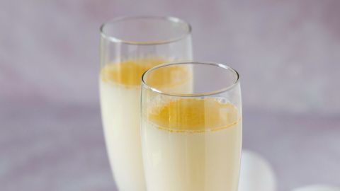 Zwei Gläser Sekt mit Eierlikör blubbern sprudelnd, umgeben von Eiern auf einem sanften rosa Hintergrund.