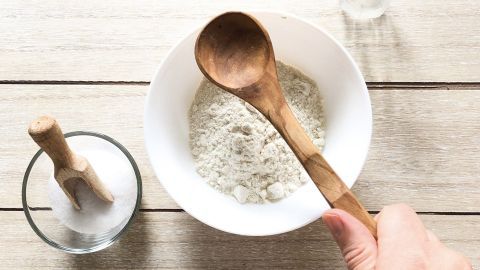 Eine hand hält einen Holzlöffel, der über eine Schüssel mit Trockenshampoo-Pulver schwenkt, auf einem hellen Tisch.
