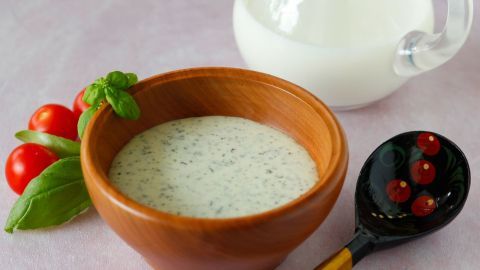 Ein Holzschälchen mit Salatdressing steht auf einem Tisch, umgeben von Kirschtomaten und einer Kanne Milch.