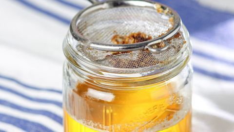 Ein durchsichtiger Glasbehälter mit Butterschmalz steht auf einem gestreiften Tuch, während ein Sieb das geschmolzene Fett filtert.