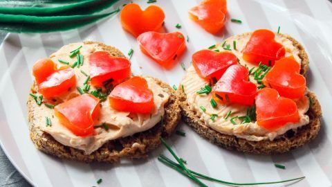 Auf einem Teller liegen zwei Pumpernickel-Herz-Taler, belegt mit Frischkäse und Tomaten in Herzform.