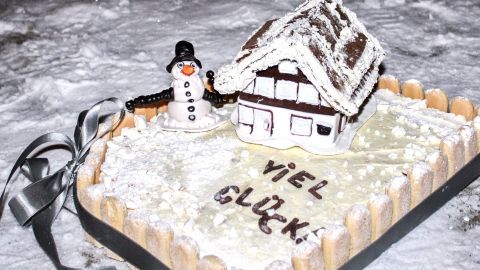 Schon beim Hausbau sollte man an mögliche Überschwemmungen denken und daher vorsorglich planen. So gehört in jeden Raum des Erdgeschosses ein schwimmender Estrich. Ein Tipp zum Schmunzeln ;-) Ein schön dekorierter Kuchen mit einem kleinen Häuschen und einem Schneemann, umgeben von Schnee, steht festlich auf einem Tisch. Der Text „VIEL GLÜCK“ ist darauf geschrieben.