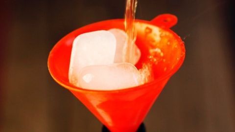 Ein roter Trichter mit Eiswürfeln steht auf einer Bierflasche, während Flüssigkeit hineingegossen wird.
