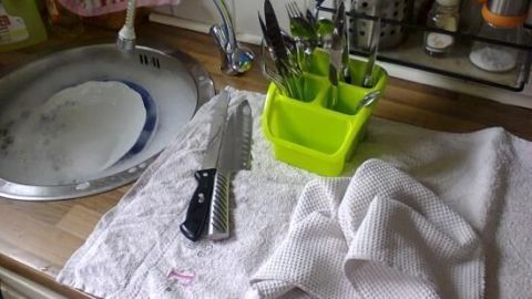 Auf einer Küchentheke liegt ein Handtuch, während Geschirrspülen im Hintergrund mit einem Waschbecken und Besteckhalter passiert.