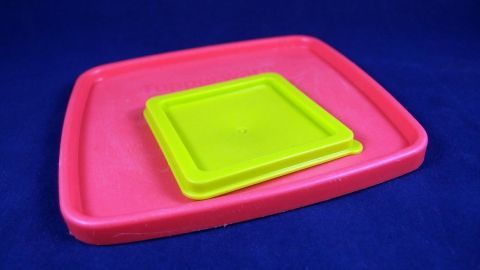 Ein rosa Tupperdeckel liegt offen auf einem blauen Hintergrund, während ein kleiner, grüner Silikondeckel darauf platziert ist.