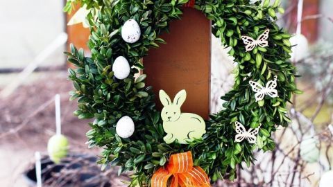 Ein Osterkranz mit einem Hasenmotiv hängt an einem Holzpfosten, dekoriert mit Ostereiern und Schmetterlingen im Garten.
