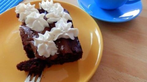 Ein Stück cremiger, schokoladiger Kuchen liegt auf einem gelben Teller, garniert mit Sahne, neben einer blauen Tasse.