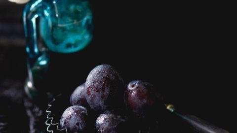 Ein blauer Glasflakon steht neben einer kleinen, schwarzen Schale mit saftigen Weinbrandpflaumen auf einem dunklen Tisch.