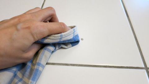 Eine Hand mit einem Tuch reinigt Wachsflecken von weißen Fliesen in einem sauberen Raum.