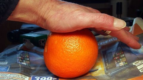 Mit diesem Tipp lassen sich Apfelsinen und auch Mandarinen leichter schälen. Eine Hand drückt eine Apfelsine auf einer Unterlage, um das Schälen zu erleichtern, während weitere Küchenutensilien sichtbar sind.