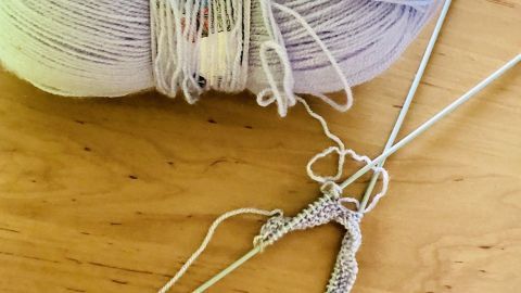 Ein Knäuel Wolle liegt auf einem Tisch, während Stricknadeln mit beginnendem Strickmesser im Vordergrund arbeiten.