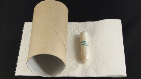 Eine Klopapierrolle liegt nebeneinander mit einem kleinen Objekt auf einem Stück Papier. Der Hintergrund ist schwarz.
