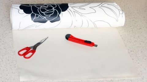 Ein Rollenstück Tapete liegt auf einer Arbeitsfläche. Neben ihm liegen eine Schere und ein Cutter, bereit zum Zuschneiden.