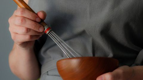 Eine Person mit einer Schürze aus Wachstuch rührt mit einem Schneebesen in einer Holzschüssel.