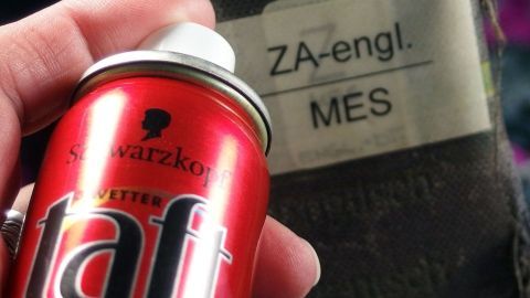 Eine rote Haarspraydose von Schwarzkopf Taft wird von einer Hand gehalten. Im Hintergrund ist ein Etikett mit "ZA-engl. MES".