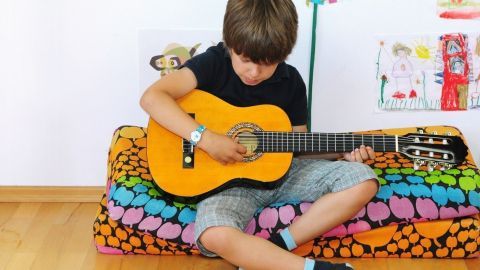 Ein Junge sitzt auf einer bunten Couch und übt konzentriert auf seiner Gitarre, um Musikinstrumente zu erlernen.