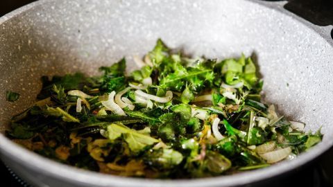 Grüner Brennnesselspinat wird in einer Pfanne mit Zwiebeln angebraten, umgeben von frischen Tomaten im Hintergrund.