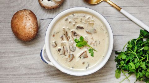 Eine cremige Champignoncremesuppe in einer weißen Schüssel, umgeben von frischen Pilzen und Petersilie auf einer Holzoberfläche.