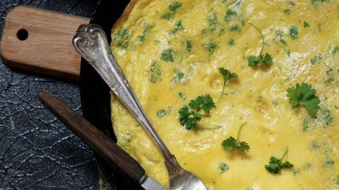 Omelette in Pfanne, bestreut mit Petersilie, liegt auf einem rustikalen Untergrund mit rustikalem Messer und Gabel.
