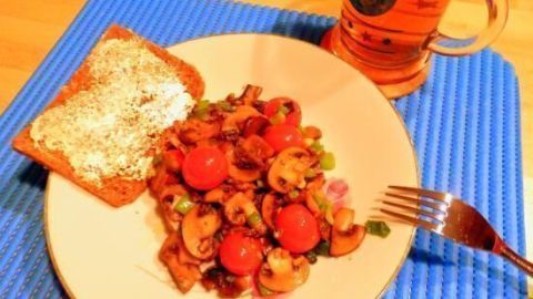 Eine bunte Pilzpfanne mit Tomaten wird auf einem Teller serviert, begleitet von einer Brotscheibe und einem Bier.