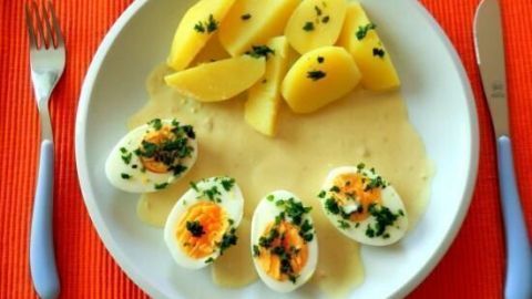 Gekochte Eier mit Senfsauce und gehackten Kräutern neben Kartoffelspalten auf weißem Teller, auf oranger Unterlage serviert.