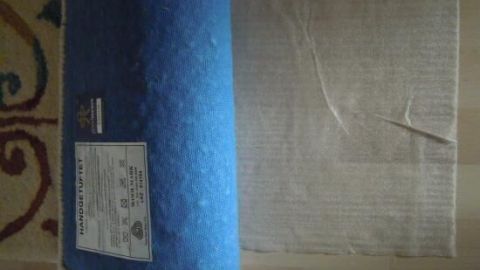 Ein Teppich liegt auf einem Holzboden, die Kante ist aufgerollt. Auf dem Rücken steht: "HANDGEKNÜPFT" mit Pflegehinweisen.