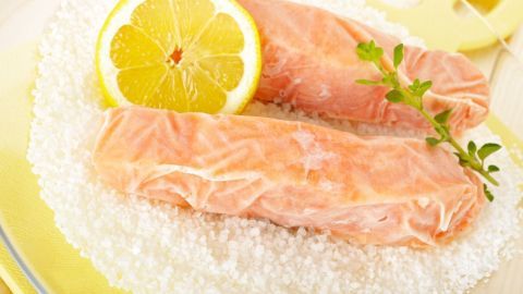 Zwei gefrorene Lachsfilets liegen auf einem Bett aus grobem Salz, garniert mit einer Zitronenscheibe und Kräutern.