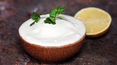 Eine cremige Salatsoße steht in einer Schale und wird von einem Zitronenstück begleitet, auf einer rustikalen Oberfläche.