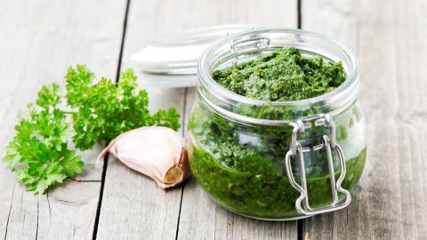 Das Glas mit dem Knoblauch-Petersilien-Pesto steht auf einem Holztisch, umgeben von frischer Petersilie und einer Knoblauchzehe.